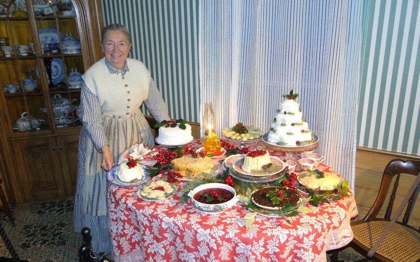 Woman in dress and apron stands next to table with many holiday foods on it
