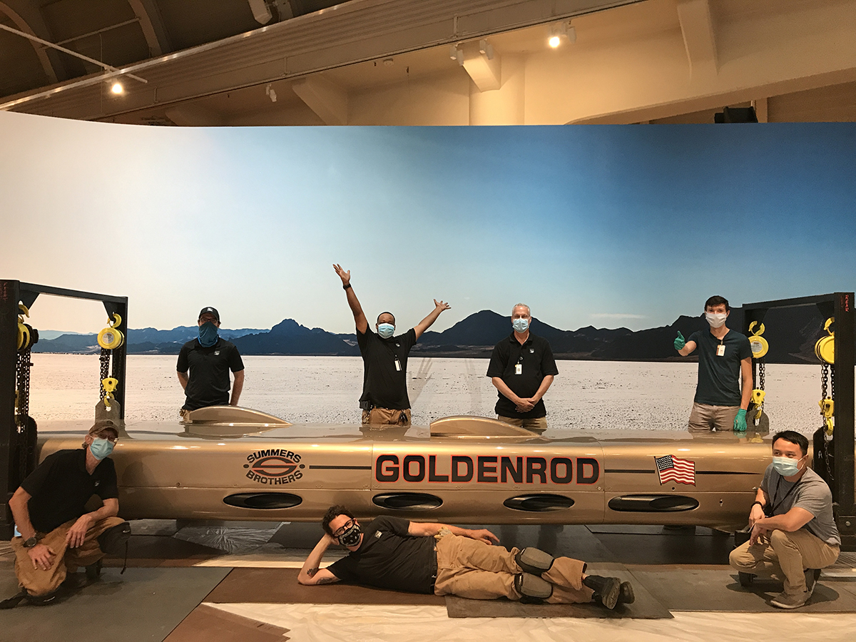 Men wearing facemasks, one with arms raised triumphantly and one giving a thumbs-up, stand, kneel, and lie around a long, low, torpedo-shaped golden car in front of a backdrop of a salt flat, mountains, and blue sky