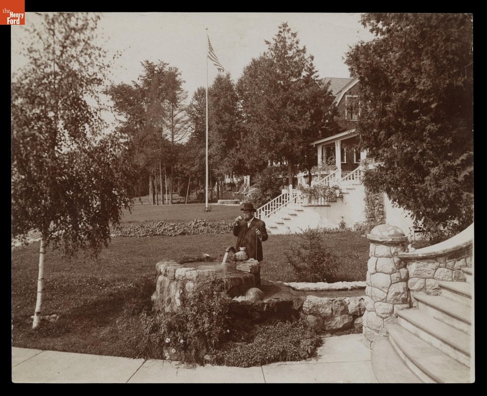 Black-and-white photo of man in suit drinking from stone well on a lawn near houses or other buildings