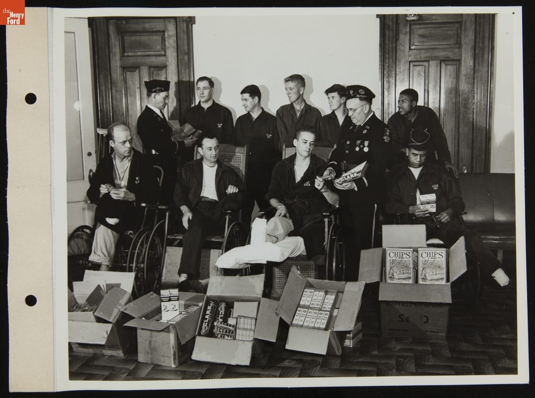 Group of men in robes and pajamas, some in wheelchairs, are handed items by two soldiers; additional items in boxes in front of the group