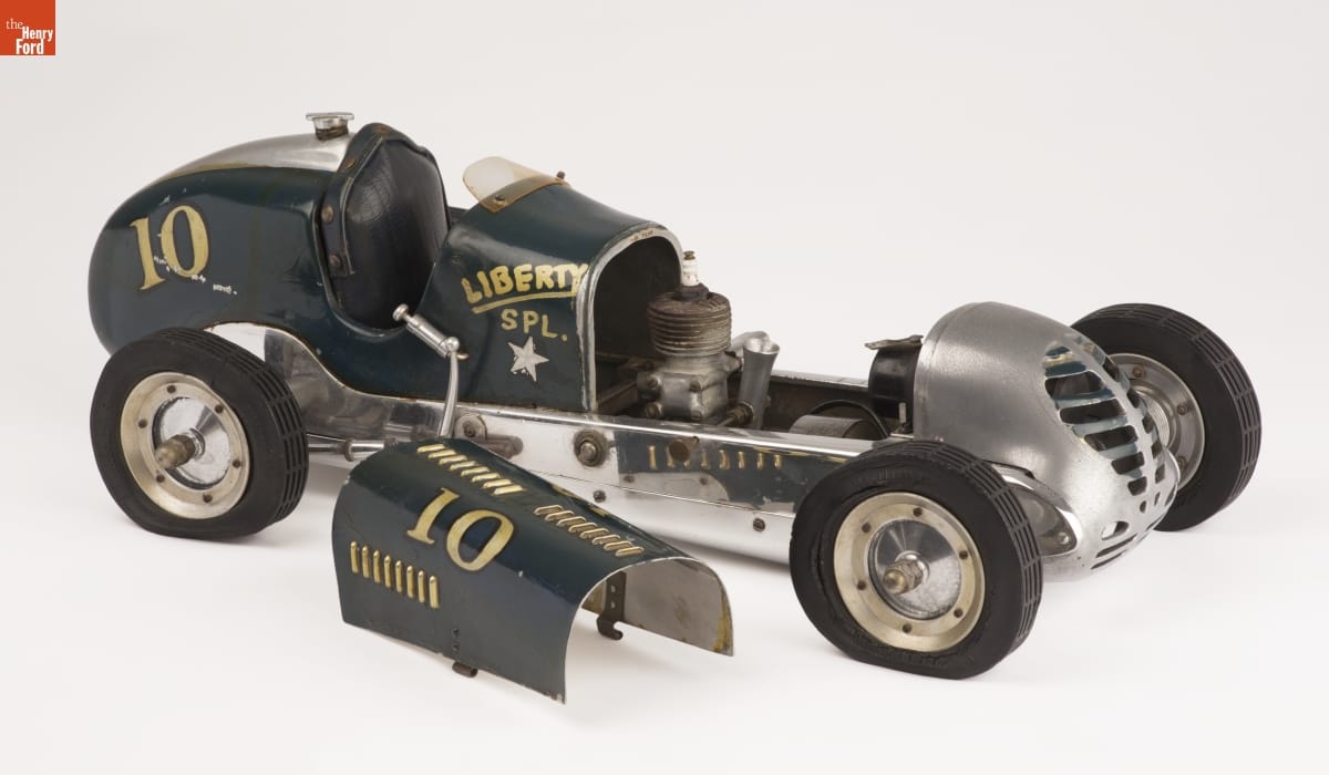 Long green model car with text and numbers on side, hood removed and sitting next to car