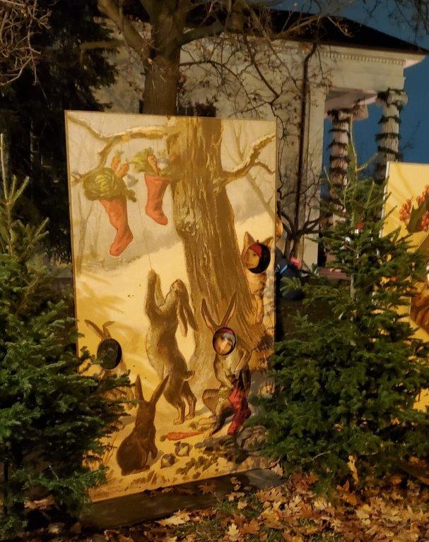 Two people stick their faces through holes in a life-size holiday postcard among evergreens