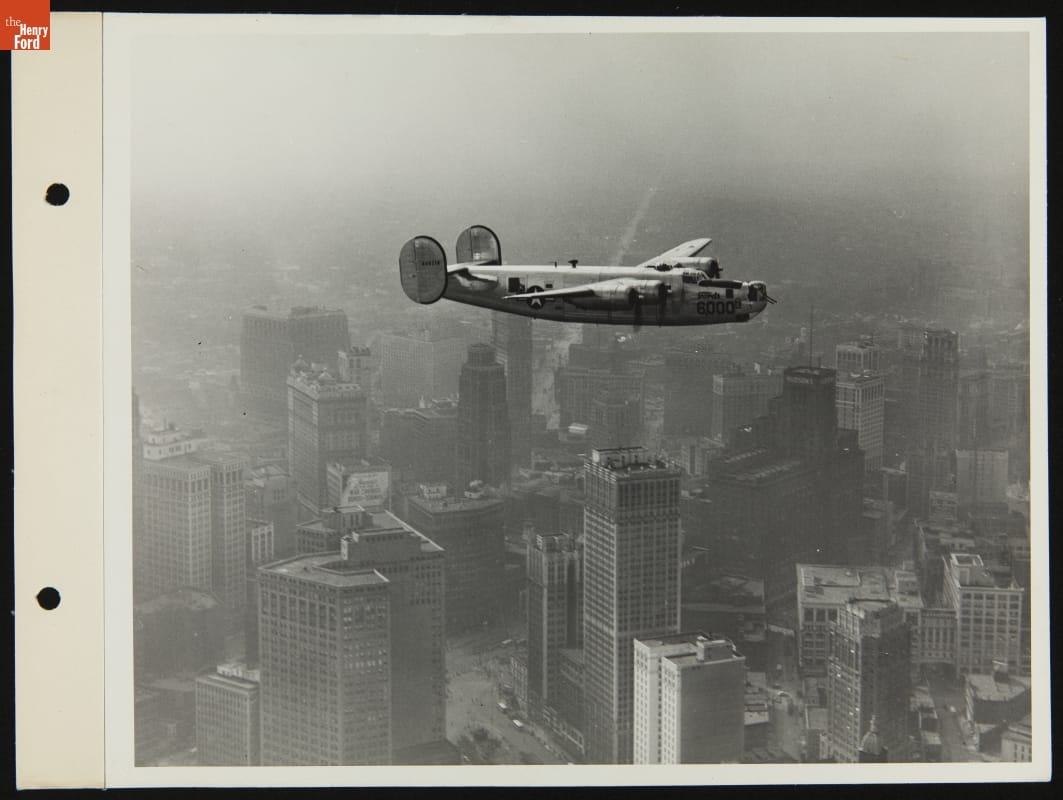 Black-and-white image of airplane flying over tall buildings.