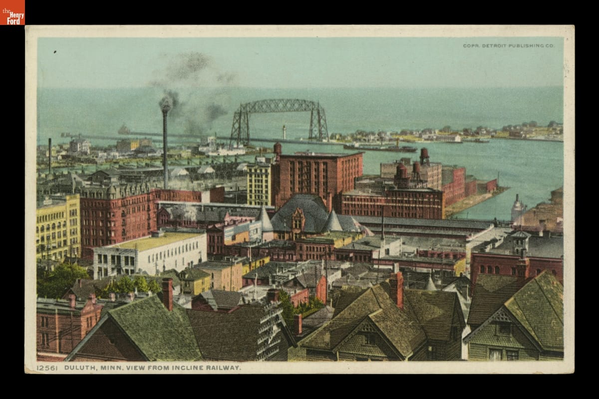 Aerial view of what appear to be houses, offices, and industrial buildings near a body of water, with smoke billowing from a smokestack
