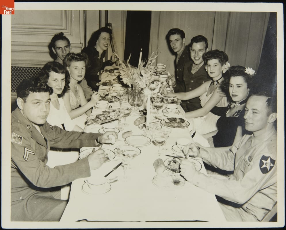 Group of men in military uniforms and women in dresses sit at a long dinner table