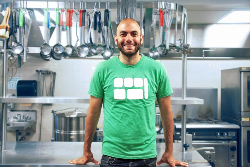 Chef Dan Giusti, founder of Brigaid Man in green t-shirt with white graphic leans against a stainless steel table in an industrial kitchen