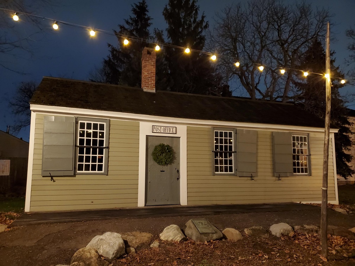 Small, beige, one-story wooden building with wreath on door and lights strung above it