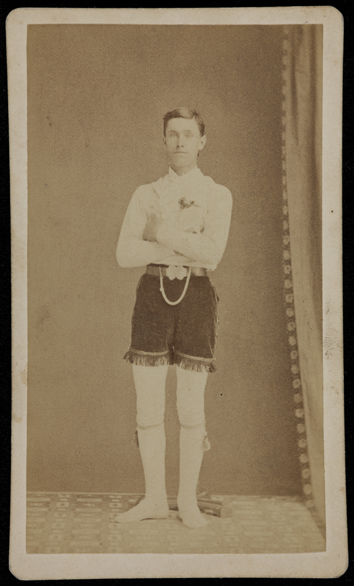 Fireman for the Alert Fire Company, Big Rapids, Michigan, 1877 Black-and-white photo of man standing with arms crossed, wearing white long-sleeved shirt, white tights (?), and dark velvet shorts with fringe