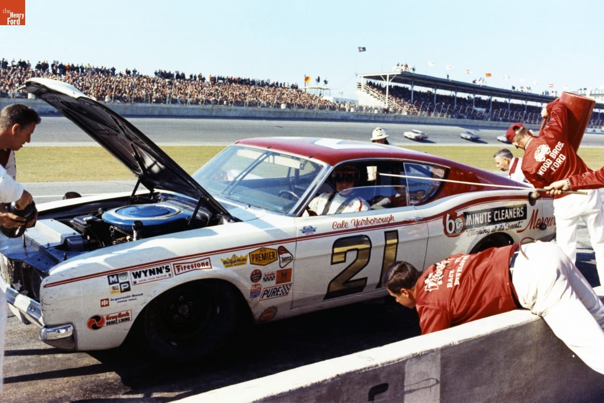 Man in helmet sits in white car with number "21" and logos on side at the side of a racetrack while several people work on the car