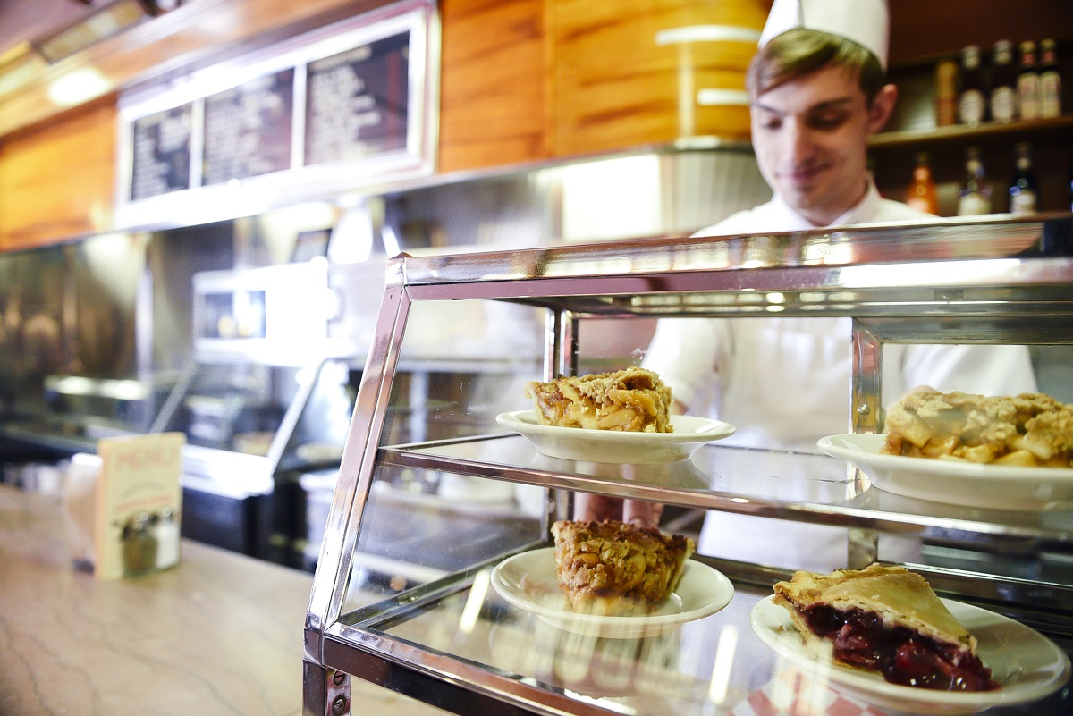Slices of pie wait for customers in Lamy's Diner A glass case filled with slices of pie sits on a lunch counter with a person in a white cap and and apron behind the counter